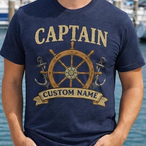 May include: Navy blue t-shirt with a nautical design. The shirt features the word "CAPTAIN" above a ship's wheel, anchors, and a banner that reads "CUSTOM NAME". The person is wearing sunglasses and blue jeans.