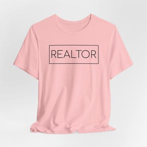 May include: A light pink t-shirt with the word "REALTOR" printed in a black rectangular box. The shirt has short sleeves and a crew neck. The text is centered on the front of the shirt.
