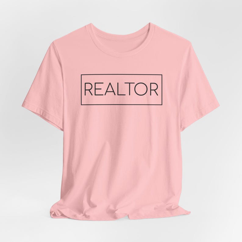 May include: A light pink t-shirt with the word "REALTOR" printed in a black rectangular box. The shirt has short sleeves and a crew neck. The text is centered on the front of the shirt.