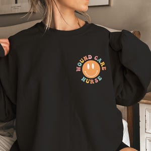Puede incluir: Sudadera negra con un gráfico de arcoíris que dice "Wound Care Nurse" con una carita sonriente en el medio.