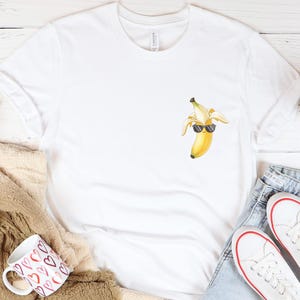 May include: White t-shirt with a watercolor illustration of a banana wearing sunglasses. The shirt is displayed with a pair of white sneakers, blue jeans, and a coffee mug with a heart pattern.