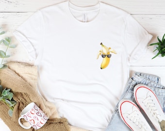 May include: White t-shirt with a watercolor illustration of a banana wearing sunglasses. The shirt is displayed with a pair of white sneakers, blue jeans, and a coffee mug with a heart pattern.