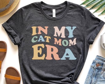 May include: Dark gray t-shirt with the text "IN MY CAT MOM ERA" in pastel colors. The shirt is a crew neck with rolled-up sleeves. The image also shows sandals, a straw hat, and denim shorts.