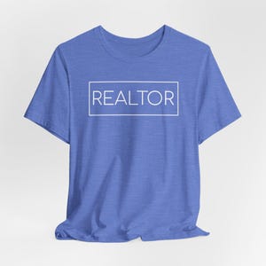 May include: A blue t-shirt with the word "REALTOR" in a white rectangular box. The shirt is made of a soft material and has short sleeves. The text is centered on the front of the shirt.
