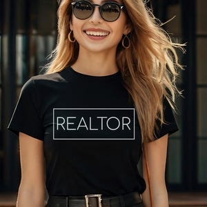 May include: Black t-shirt with the word "REALTOR" in a white rectangular box. The person is wearing sunglasses and hoop earrings. The shirt has short sleeves and a crew neck. The person is smiling.