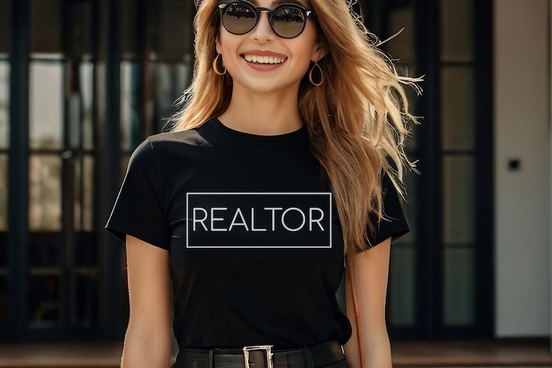May include: Black t-shirt with the word "REALTOR" in a white rectangular box. The person is wearing sunglasses and hoop earrings. The shirt has short sleeves and a crew neck. The person is smiling.