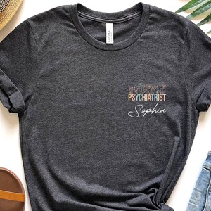 May include: A dark heather grey t-shirt with the text "PSYCHIATRIST" in rainbow colors and the name "Sophia" in white script.