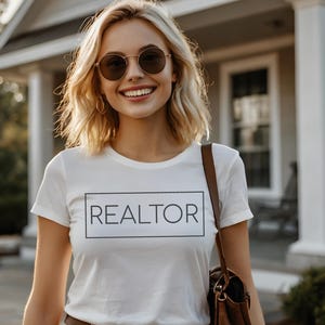 May include: A white t-shirt with the word "REALTOR" in a black rectangular box. The person is wearing sunglasses and a brown shoulder bag. The background includes a house and greenery.