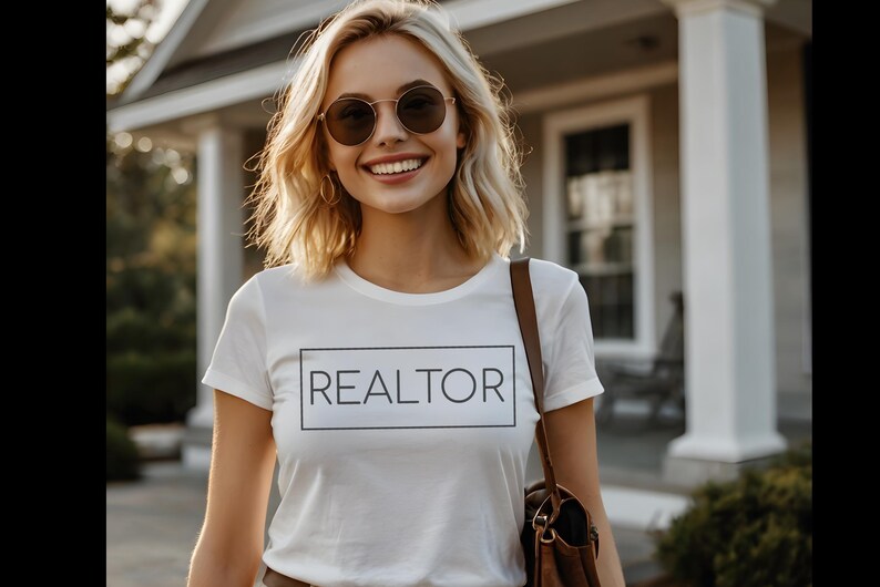 May include: A white t-shirt with the word "REALTOR" in a black rectangular box. The person is wearing sunglasses and a brown shoulder bag. The background includes a house and greenery.