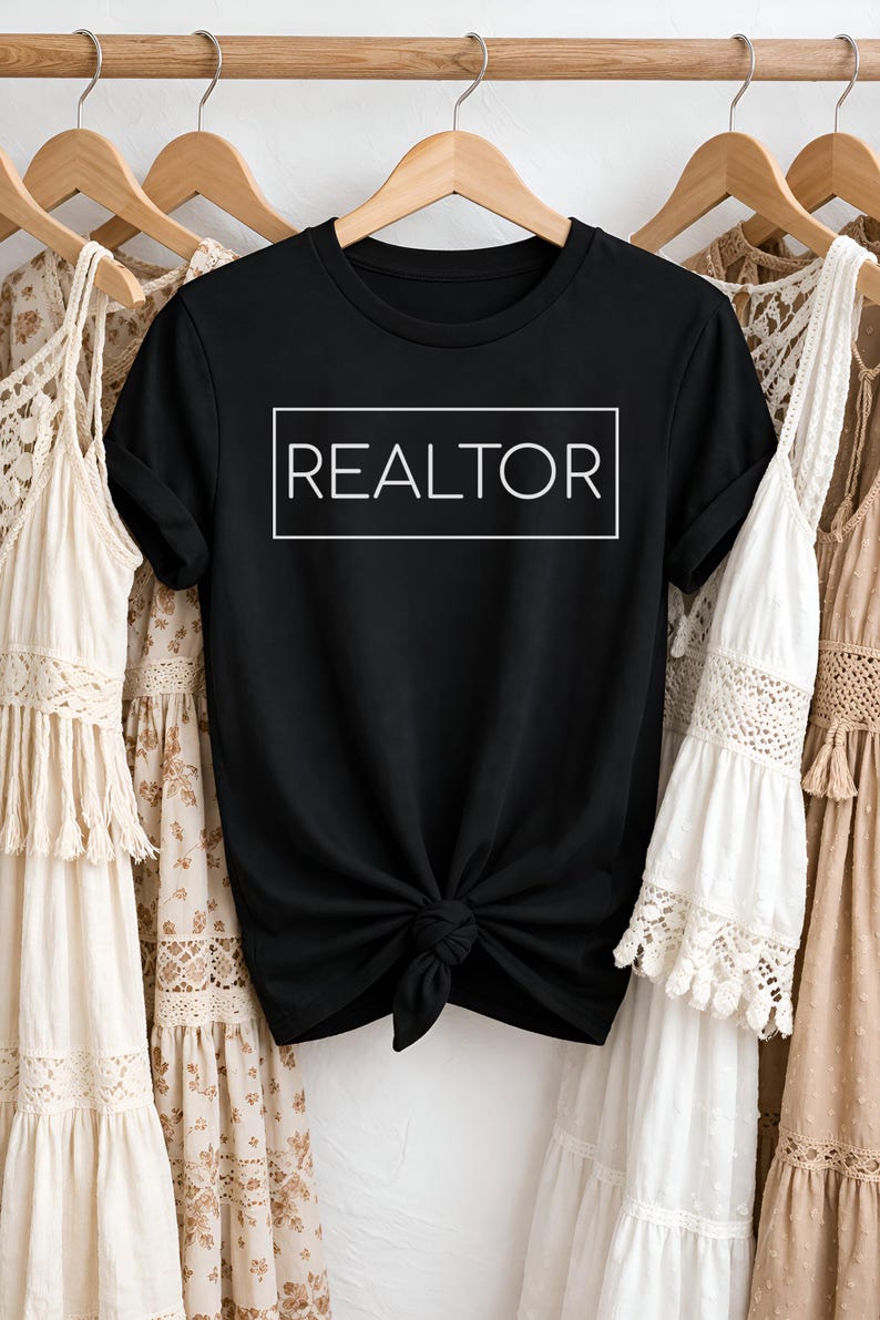 May include: Black t-shirt with the word "REALTOR" in a white rectangular box. The shirt is knotted at the waist and hangs on a wooden hanger. Other clothing items hang on adjacent hangers.