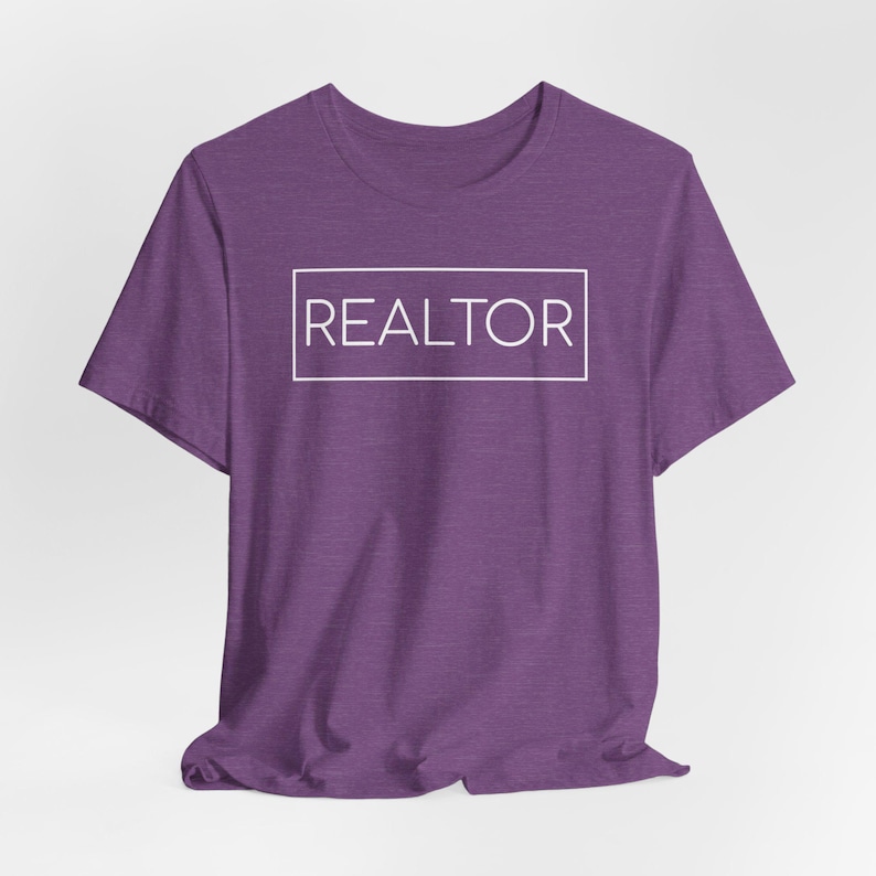 May include: A purple t-shirt with the word "REALTOR" in a white rectangular box. The shirt has short sleeves and a crew neck. The fabric has a heathered texture.