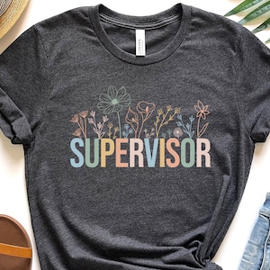 May include: A dark grey t-shirt with a floral design and the word "SUPERVISOR" in rainbow colours.