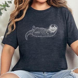 May include: Dark gray t-shirt featuring a white line drawing of a relaxed otter floating on its back. The otter has a detailed face and is positioned horizontally across the shirt. The shirt is made of a soft material.
