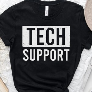 May include: Black t-shirt featuring the text "TECH SUPPORT" in a white, distressed font within a white rectangle. The shirt is shown on a white surface with a hat, a book, and a coffee cup.