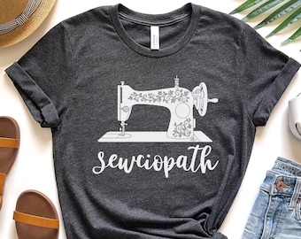 May include: Dark gray t-shirt featuring a white illustration of a vintage sewing machine with floral details. The word "sewciopath" is written in cursive below the machine. The shirt is a casual wear item.