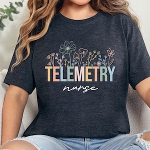 May include: Dark gray t-shirt with the words "TELEMETRY nurse" in colorful block letters and a floral design above. The shirt is a crew neck and has short sleeves.