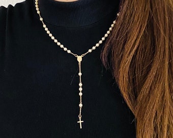 Puede incluir: Collar de rosario dorado con pequeñas cuentas blancas redondas. El collar tiene un colgante de cruz y un medallón central. La cadena está colocada sobre un cuello alto negro.
