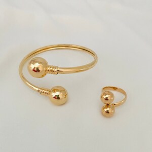 May include: A gold-colored jewelry set featuring a cuff bracelet and a ring. The bracelet has two large, spherical accents and a spiral design. The ring also has two spherical accents. The jewelry is displayed on a white surface.