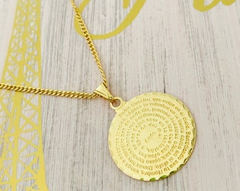 May include: A gold necklace with a round pendant featuring a spiral inscription of the Lord's Prayer in Spanish. The pendant is on a gold chain.