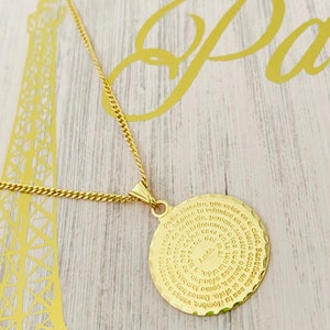 May include: A gold necklace with a round pendant featuring a spiral inscription of the Lord's Prayer in Spanish. The pendant is on a gold chain.