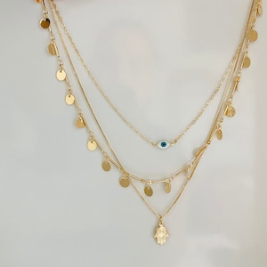 Könnte beinhalten: Eine mehrreihige goldene Halskette mit einem Hamsa-Hand-Anhänger und einem blauen Glücksbringer-Anhänger. Die Kette besteht aus mehreren Kettensträngen mit kleinen goldenen Scheiben, die an den Seiten hängen.