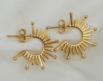 May include: A pair of gold hoop earrings with a sunburst design. The earrings are made of metal and have a circular shape with a series of small, pointed rays extending outward from the center.