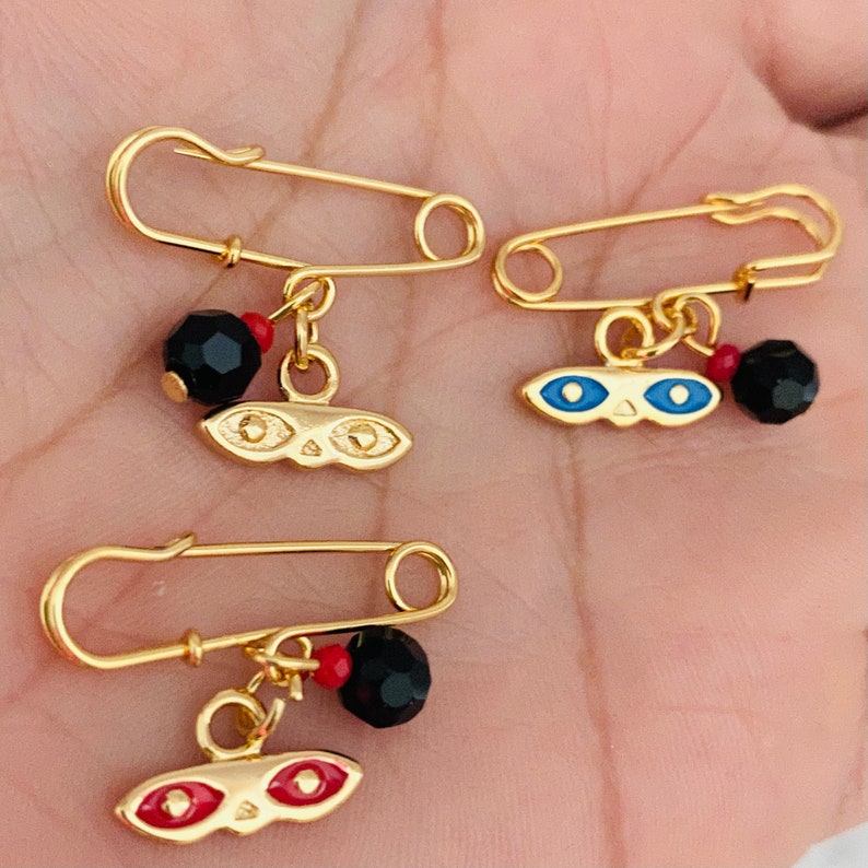 May include: Three gold safety pin charms with black and red beads and a gold eye charm. The eye charms have blue, red, and gold accents.