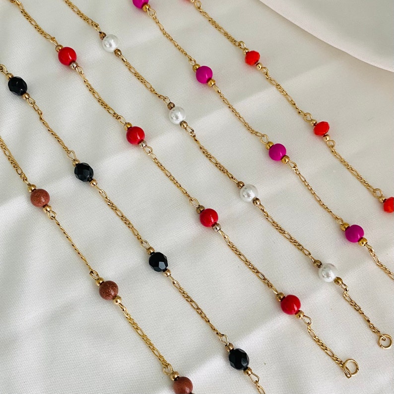 May include: A set of six gold chain necklaces with different colored beads, including red, black, white, pink, and orange. The necklaces are displayed on a white surface.
