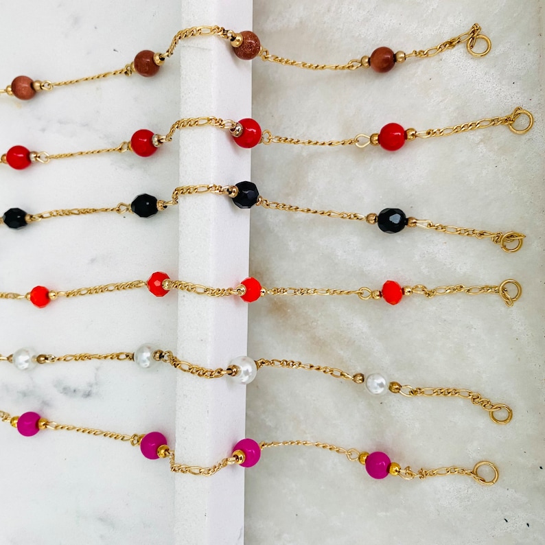 May include: Seven gold chain bracelets with different colored beads. The beads are red, black, white, orange, and pink.