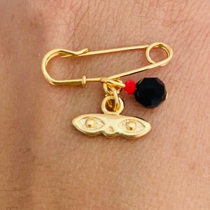 May include: A gold safety pin with a black and red bead charm and a gold mask charm. The mask has two gold circles for eyes.