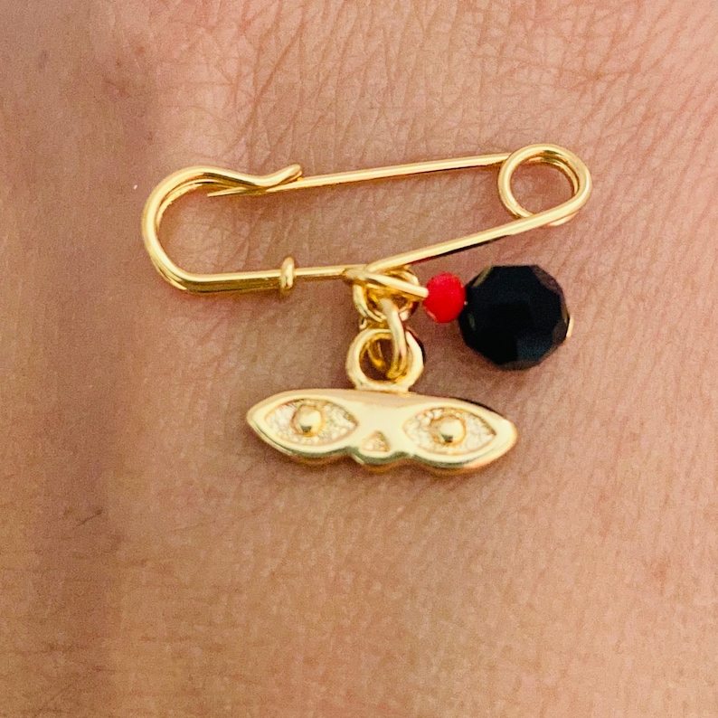 May include: A gold safety pin with a black and red bead charm and a gold mask charm. The mask has two gold circles for eyes.