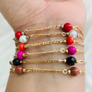 May include: A set of seven gold chain bracelets with colorful beads. Each bracelet features a different colored bead, including red, white, orange, pink, black, and brown.