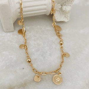 Peut inclure: Un collier en chaîne dorée avec plusieurs petits pendentifs ronds et un plus grand pendentif en forme de pièce de monnaie. La chaîne est de style délicat et lié.