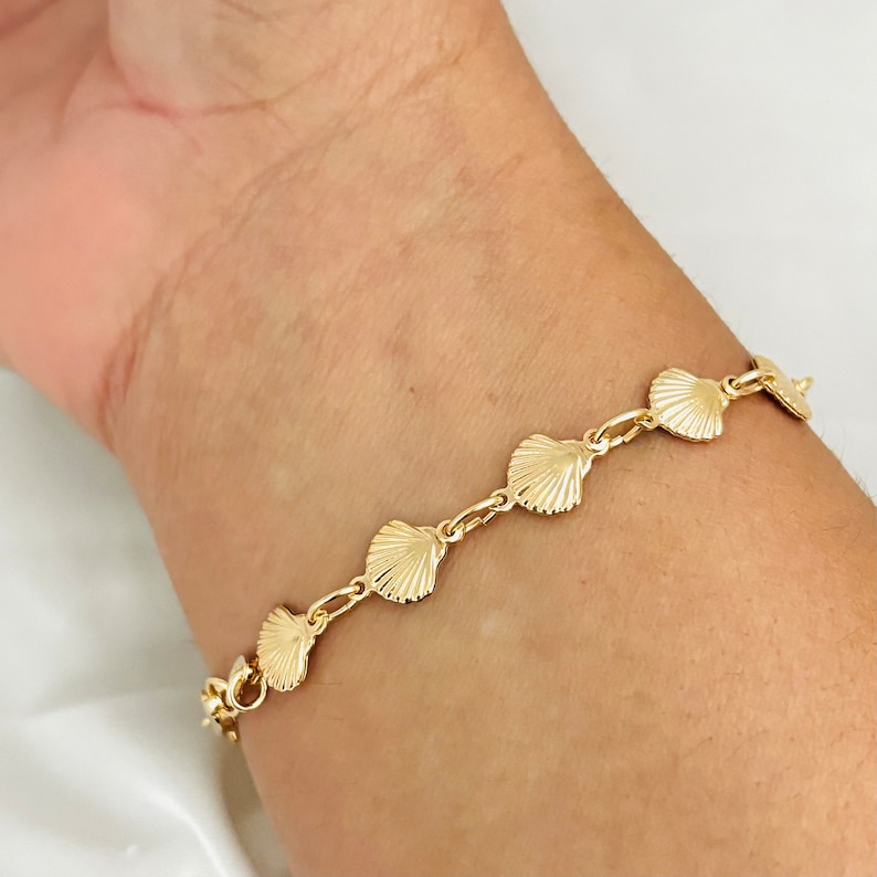 May include: A gold bracelet with a chain link design and seashell charms. The bracelet is on a person's wrist.