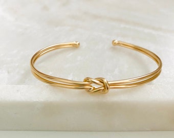 Peut inclure: Un bracelet manchette doré avec un motif de nœud. Le bracelet présente un design à double bande avec un nœud au centre. Le bracelet est fait d'un matériau métallique brillant et est présenté sur une surface blanche.
