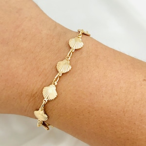 May include: A gold bracelet with a chain link design and seashell charms. The bracelet is on a person's wrist.