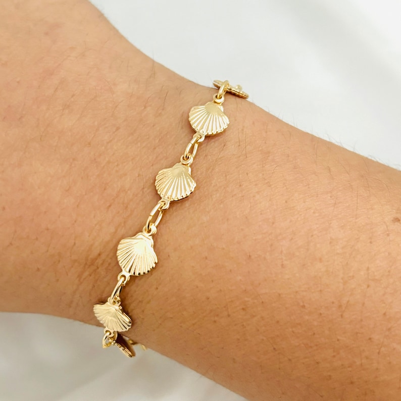 May include: A gold bracelet with a chain link design and seashell charms. The bracelet is on a person's wrist.