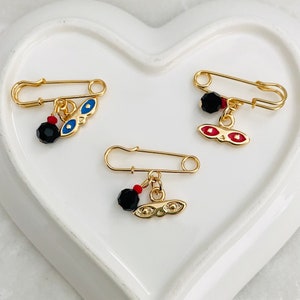 May include: Three gold safety pins with black beads and charms. The charms are gold with blue and red enamel eyes.