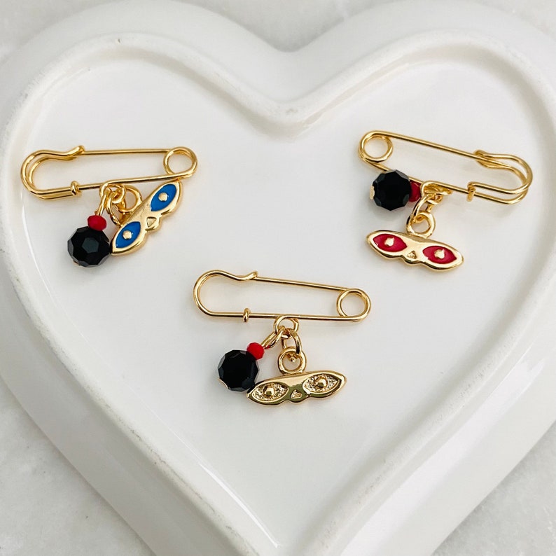 May include: Three gold safety pins with black beads and charms. The charms are gold with blue and red enamel eyes.