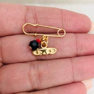 May include: A gold safety pin with a black bead and a gold charm with two eyes. The charm is attached to the pin with a gold chain.
