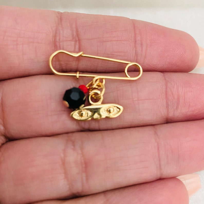 May include: A gold safety pin with a black bead and a gold charm with two eyes. The charm is attached to the pin with a gold chain.