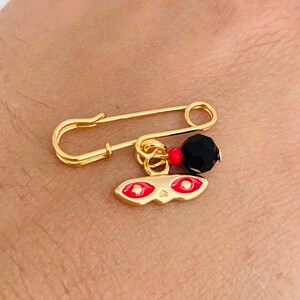 May include: A gold safety pin with a red and black charm. The charm features a mask with two red eyes.