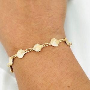 May include: A gold bracelet with a chain link design and seashell charms. The bracelet is on a person's wrist.