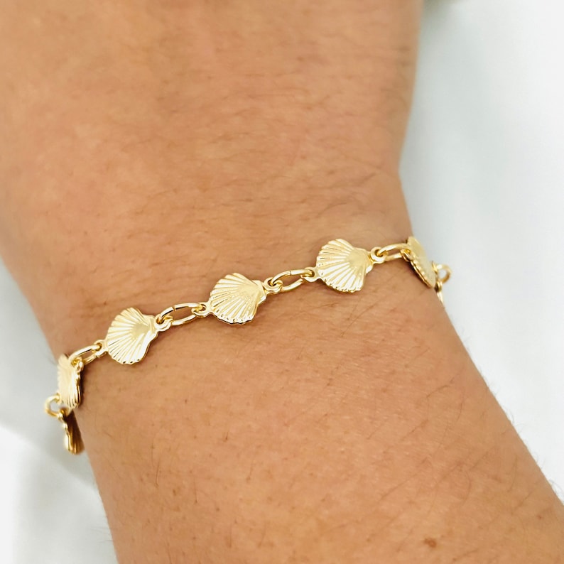May include: A gold bracelet with a chain link design and seashell charms. The bracelet is on a person's wrist.