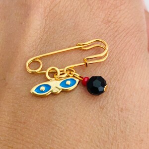 May include: A gold safety pin with a charm attached. The charm features a blue evil eye design and a black bead.