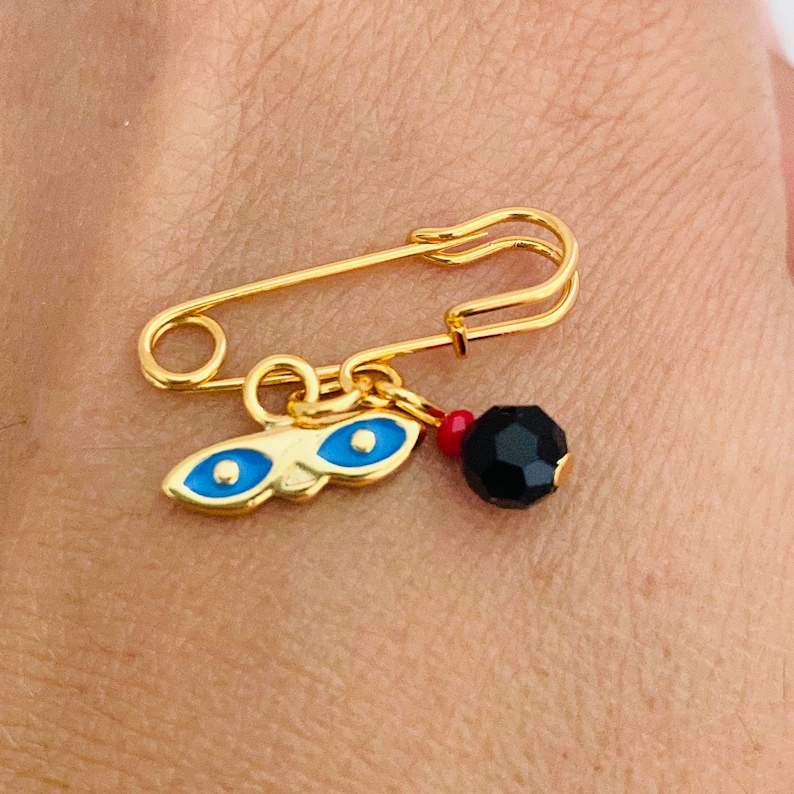 May include: A gold safety pin with a charm attached. The charm features a blue evil eye design and a black bead.