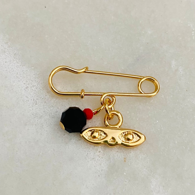 May include: A gold safety pin with a black bead and a gold charm with a face and two eyes. The charm is attached to the pin with a red bead.