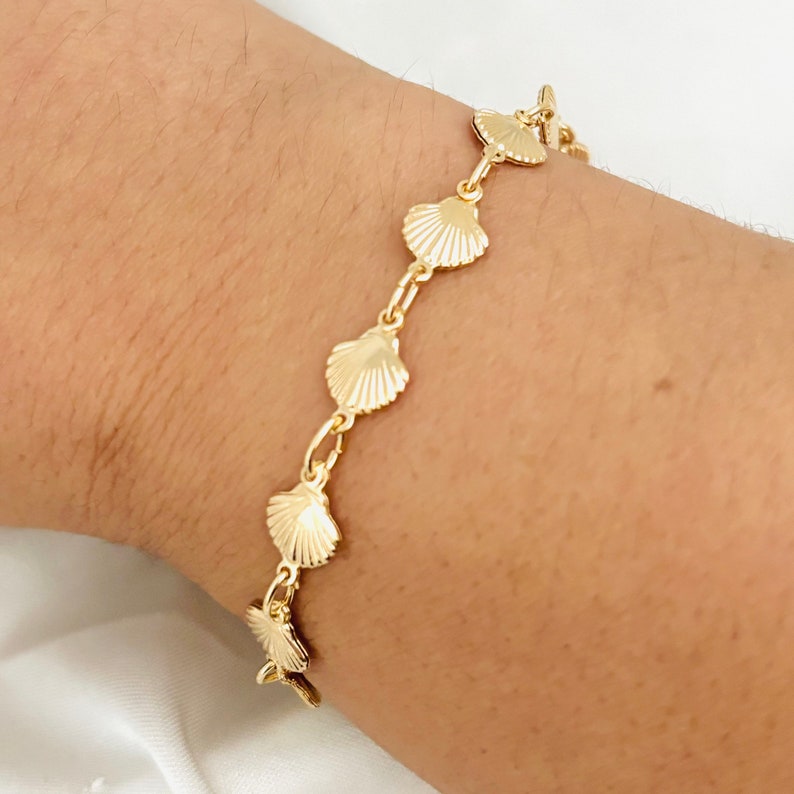 May include: A gold chain bracelet with a series of small seashell charms. The bracelet is on a person's wrist.