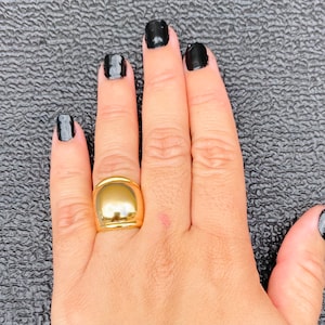 May include: A gold ring with a large, rounded, dome-shaped top. The ring is worn on the middle finger of a hand with black nail polish.