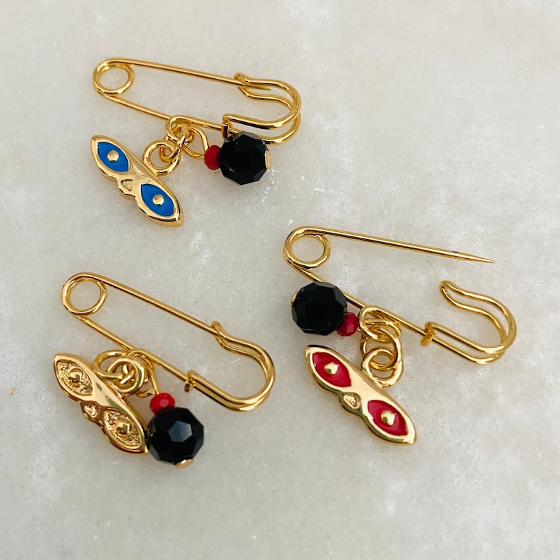 May include: Two gold safety pins with charms. One charm is a blue evil eye with red accents and the other is a red evil eye with red accents. Both charms have a black bead.